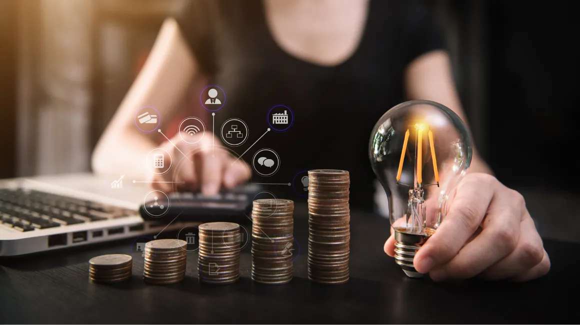 kobieta pracująca przy laptopie, trzymająca żarówkę obok stosu monet