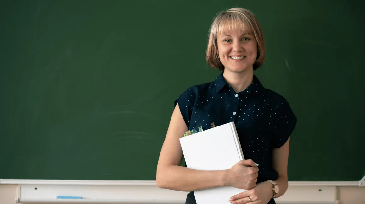 Uśmiechnięta nauczycielka stojąca przed tablicą, trzymająca w rękach segregator z dokumentami